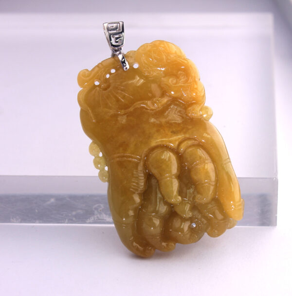 A stunning close-up of the Natural Yellow Jadeite Buddha's Hand Pendant, showcasing its rich amber-yellow hue, intricate carving details, and the elegant 18K white gold clasp. The pendant rests on a soft velvet background, highlighting its lustrous texture.