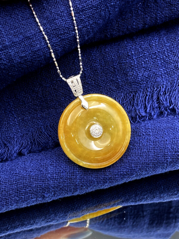 A stunning close-up of the Natural Yellow Jadeite Peace Button Pendant, showcasing its warm amber-like hue, intricate 18K white gold inlay, and sparkling diamond accents. The pendant rests elegantly on a velvet background, highlighting its luxurious craftsmanship.