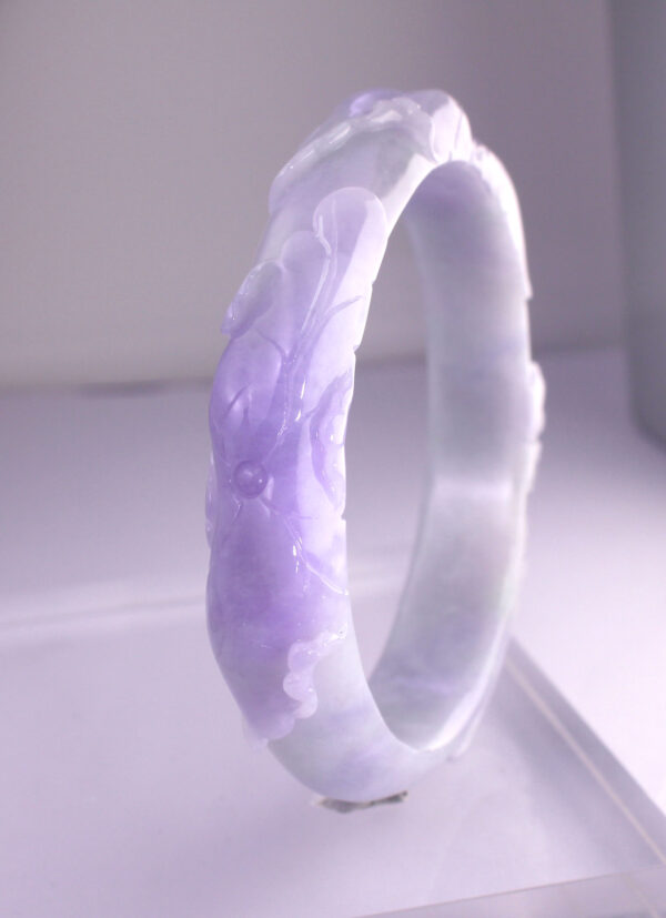 A stunning close-up of the Natural Violet Jadeite Relief Bracelet, showcasing its intricate floral relief carvings and rich violet hue against a soft background. The polished jadeite surface reflects light beautifully, highlighting its fine texture and craftsmanship.