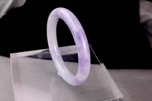 A stunning Natural Violet Jadeite Bracelet resting on a silk cloth, showcasing its serene violet hue and smooth, polished surface. The light reflects off the jadeite's delicate luster, highlighting its elegant charm and intricate craftsmanship.