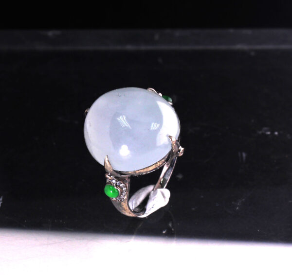 A stunning close-up of the Natural Ice White Jadeite Cabochon Ring, showcasing its smooth, translucent jadeite centerpiece set in sleek black 18K gold, accented with sparkling diamonds and vibrant green jadeite side stones.
