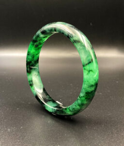 A close-up of a translucent icy jadeite bangle with vibrant green Huaqing patterns, resting on a silk cloth. The smooth polish and natural color transitions highlight its elegance and craftsmanship.