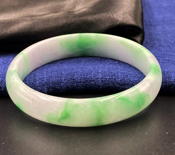 A close-up of a stunning White Base Green Jadeite Bangle, showcasing its smooth, semi-transparent texture with vibrant green patches against a pure white background. The round bangle design rests elegantly on a wrist, reflecting light beautifully.