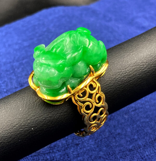 A stunning 18K yellow gold ring featuring a meticulously carved jadeite pixiu with lifelike details, set against a delicate filigree band. The vibrant green jadeite showcases a bright glassy luster, making it a luxurious and auspicious accessory.