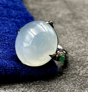 A stunning close-up of the Natural Ice White Jadeite Cabochon Ring, showcasing its smooth, translucent jadeite centerpiece set in sleek black 18K gold, accented with sparkling diamonds and vibrant green jadeite side stones.
