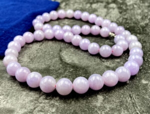 A stunning close-up of natural violet jadeite beads with a smooth, polished surface, showcasing their soft purple-to-pink gradient. The 18K white gold clasp adds a refined touch, complementing the beads' elegance.